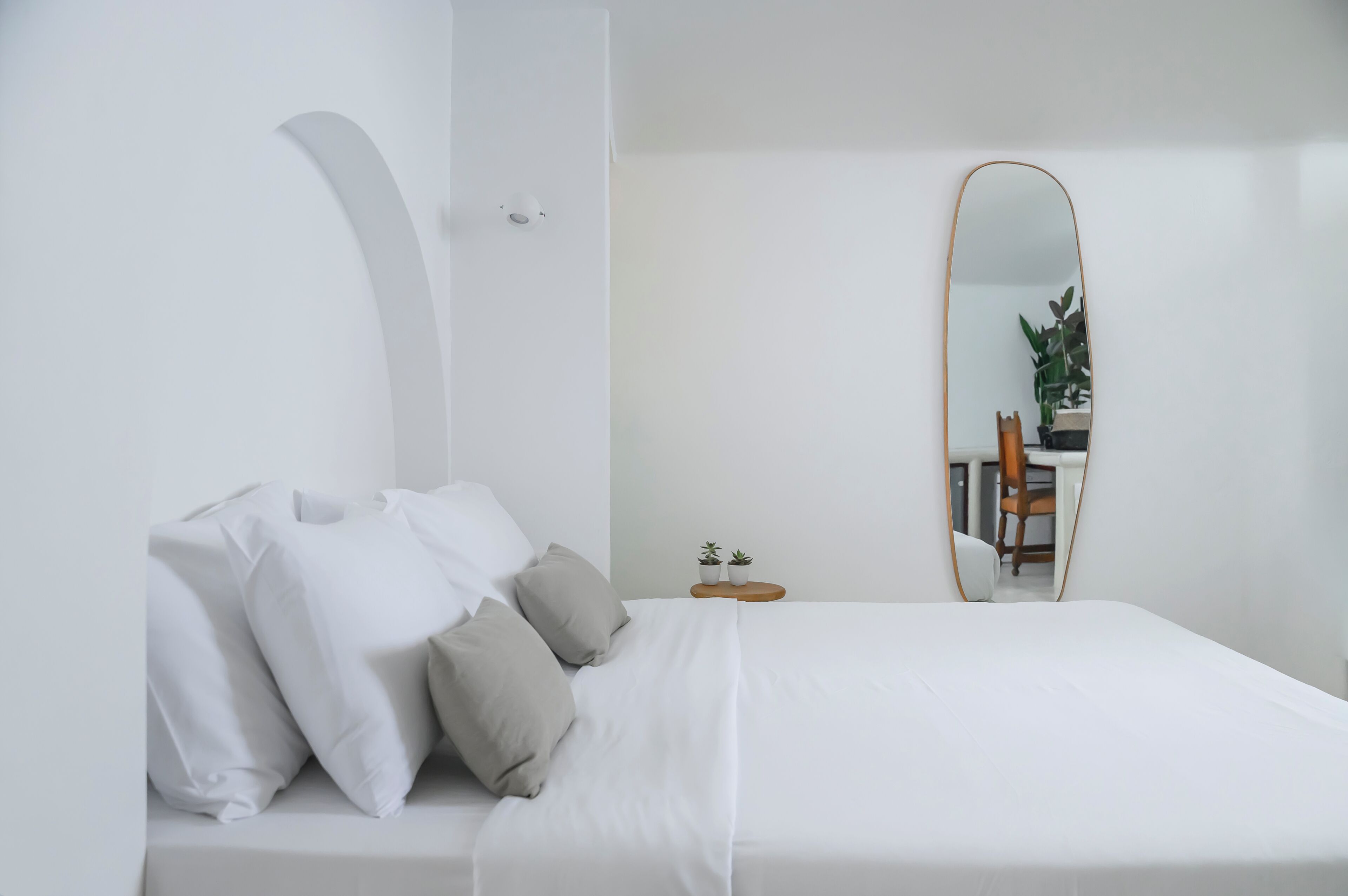 Une chambre à coucher au style minimaliste, avec un lit, une table de chevet, un miroir et une chaise à l’arrière-plan.
