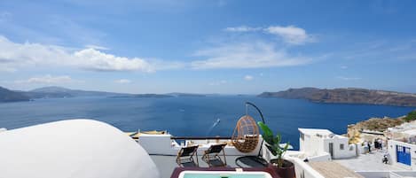 A rooftop pool with a view of the sea, white buildings, and a hanging chair.