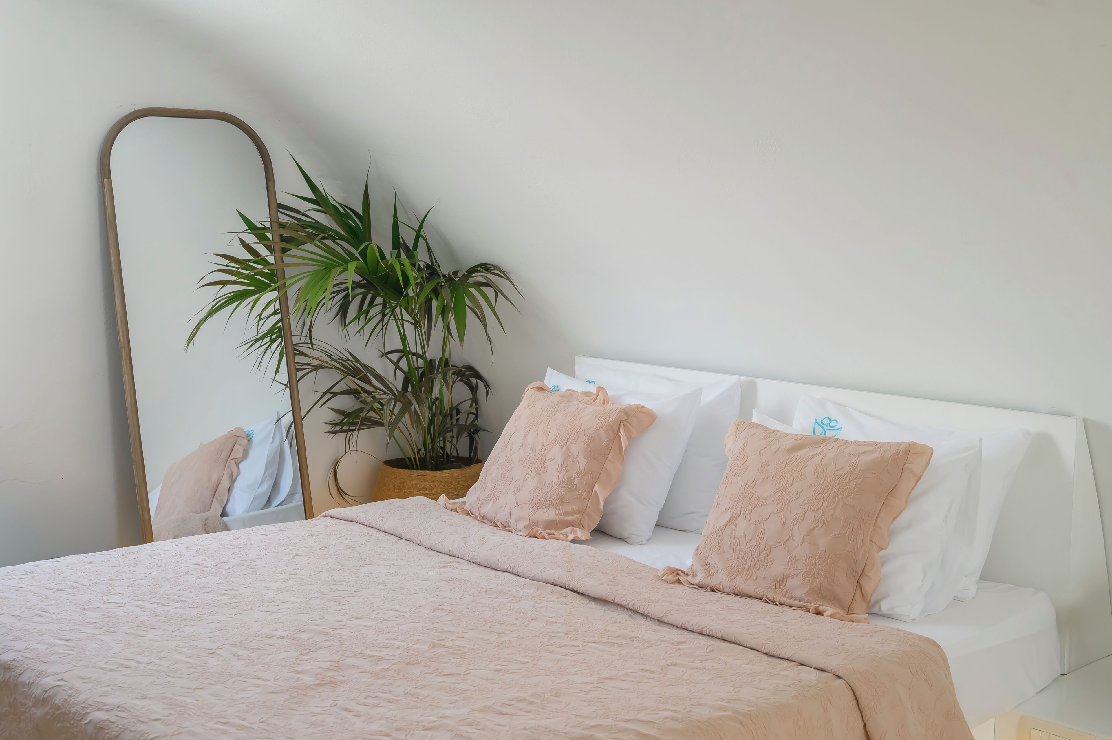 A neatly made bed with a pink blanket, white pillows, and a potted plant beside a mirror.