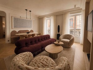 A living room with a sofa, chairs, a round coffee table, a dining table with chairs, a large framed artwork, and a television. - Madame Luxury 3BR 6P with AC (Paris)