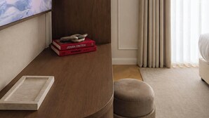 Une chambre d’hôtel moderne avec un bureau en bois, un téléviseur à écran plat, quelques livres, un pouf confortable et une grande fenêtre avec des rideaux.