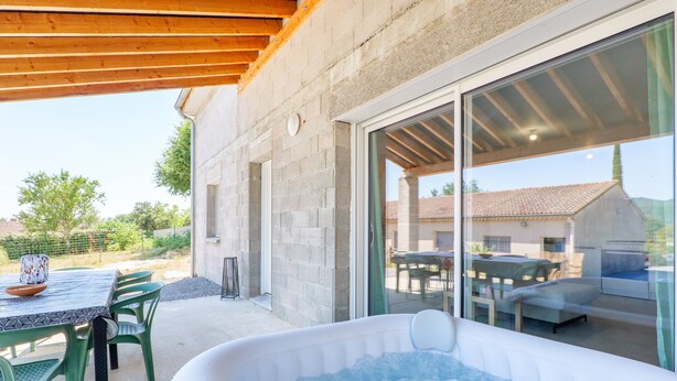 A patio area with a hot tub, outdoor seating, and a view of a house through a glass door. - Holiday Home 'Le Gecko' with Private Terrace and Wi-Fi (Saint-Germain)