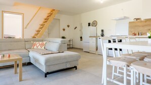 A modern living room with a sofa, a dining table with chairs, a kitchen area, and a staircase. - Holiday Home 'Le Gecko' with Private Terrace and Wi-Fi (Saint-Germain)