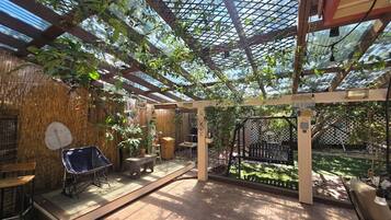 Un espace extérieur abrité, doté d’un plancher en bois, d’une table, de chaises et d’une pergola couverte de plantes grimpantes.