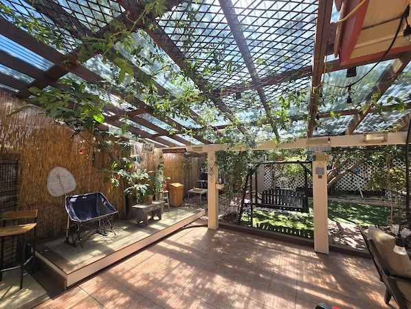 Un'area esterna coperta con pavimento in legno, un tavolo, sedie e una pergola ricoperta di piante rampicanti.