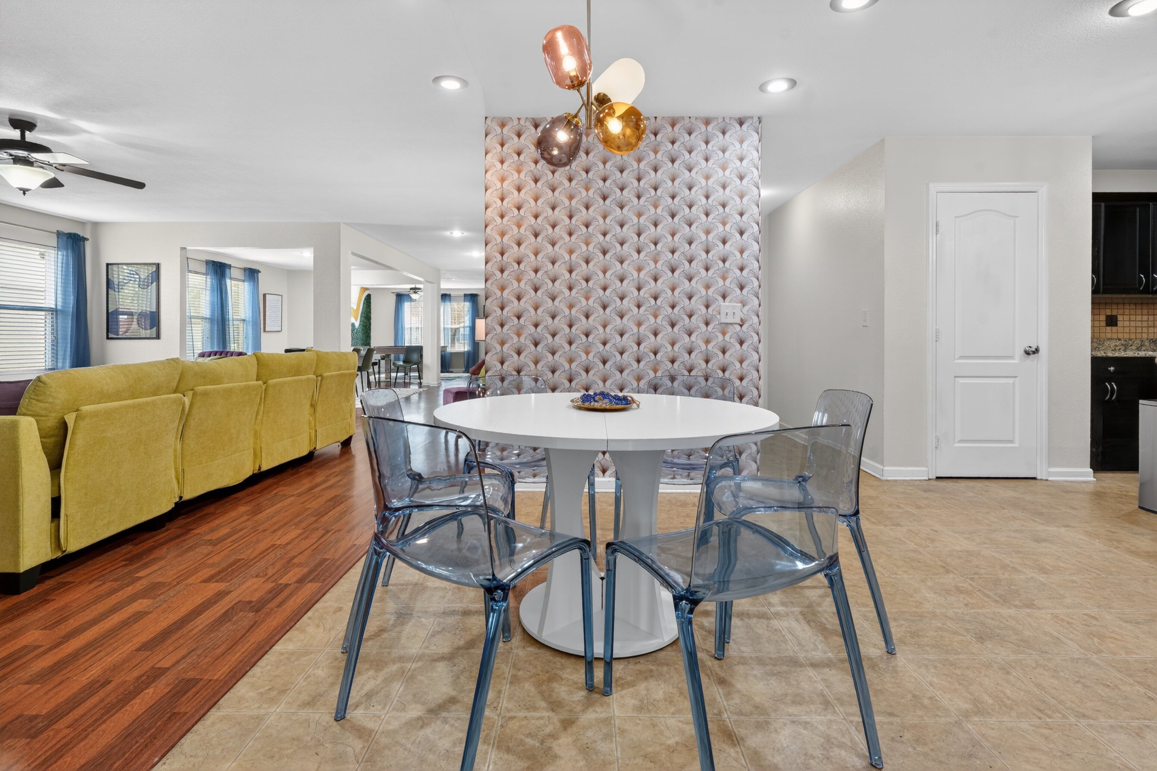 A modern living room with a yellow sofa, a round glass table with chairs, a decorative wall, and a ceiling fan.