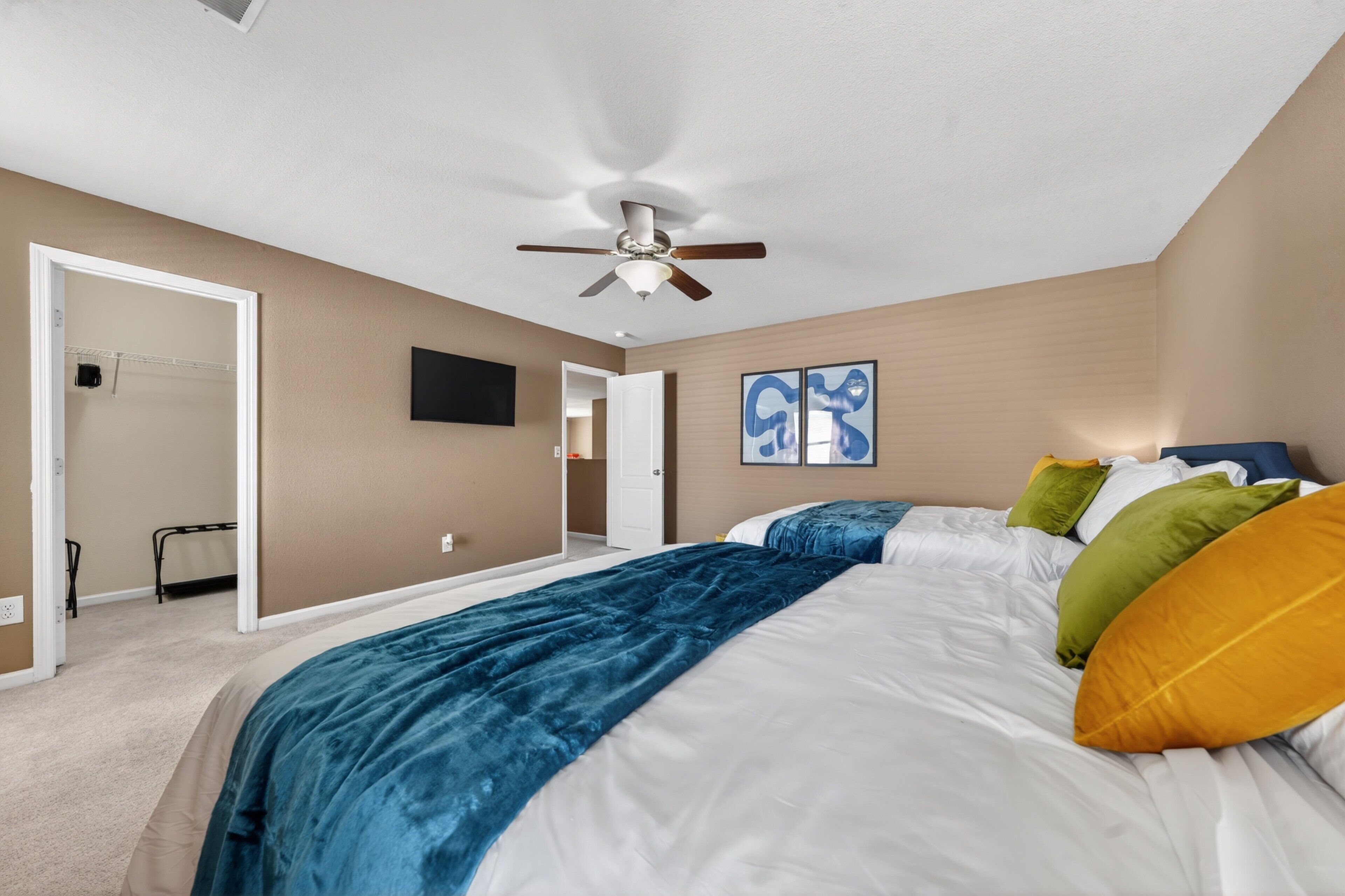 A bedroom with a bed, ceiling fan, artwork, and a door leading to another room.