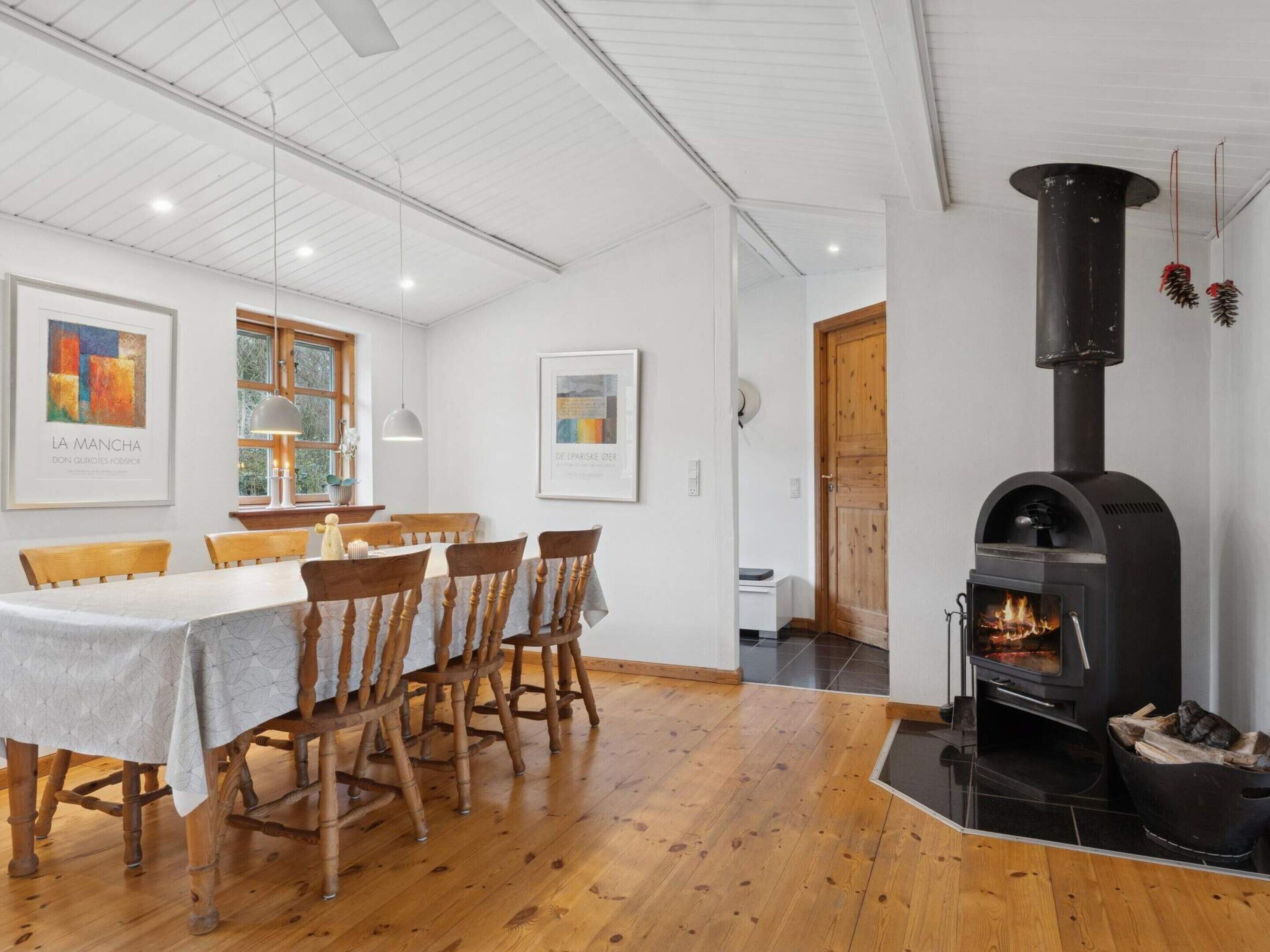 A dining area with a wooden table and chairs, a black wood stove, and framed artwork on the walls.