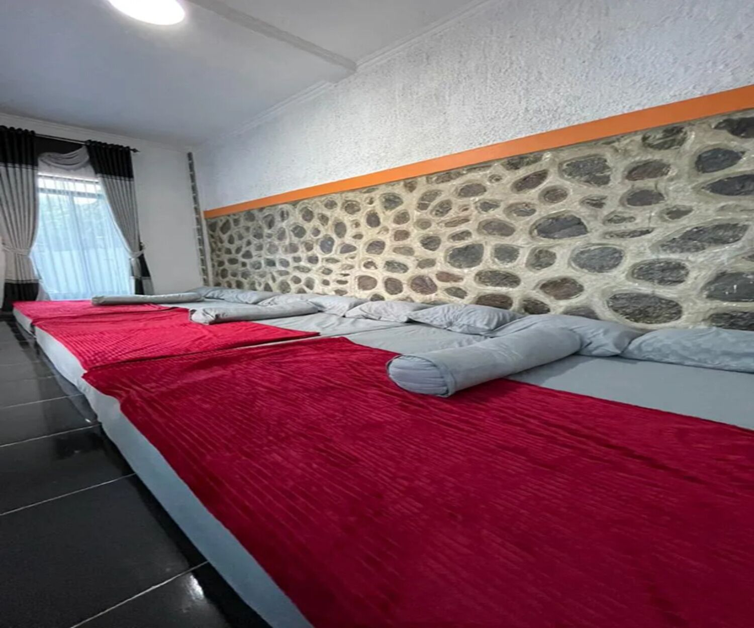 A hotel room with a stone wall headboard, a large bed with red and grey bedding, and a window with curtains.
