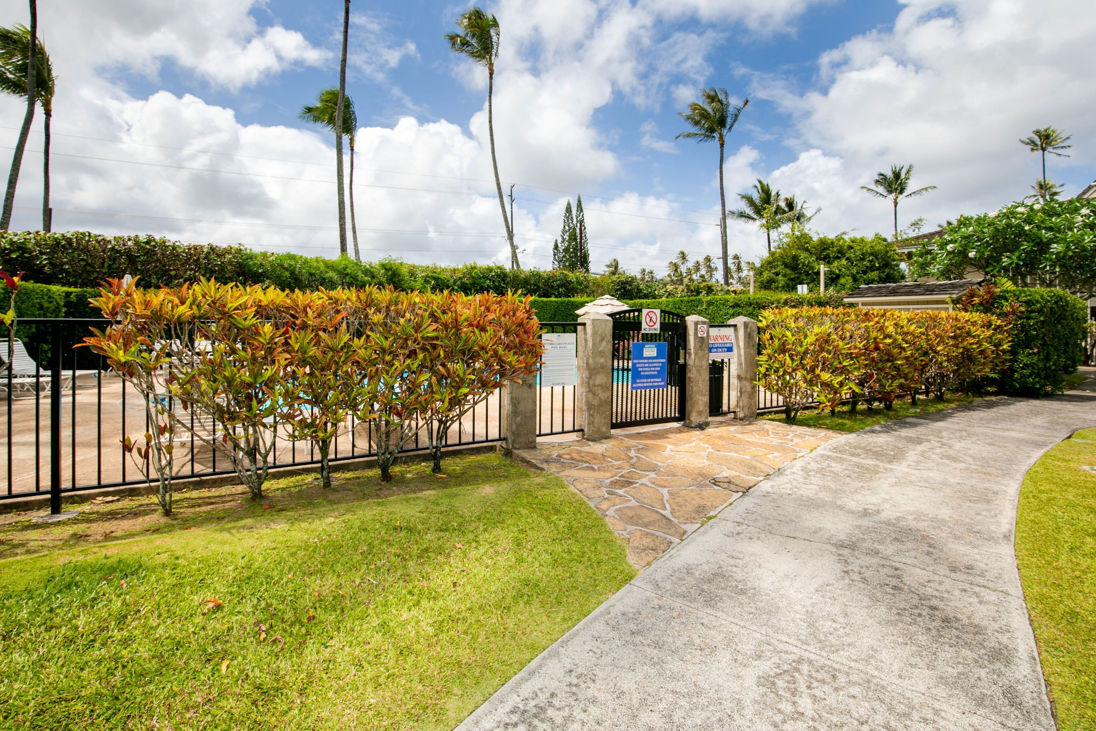 A gated entrance with a stone pathway leading to a building, surrounded by lush greenery and palm trees.