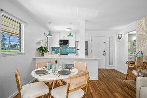 A modern kitchen with white cabinets, a central island, and a dining area with a round table and wicker chairs. - Kauai Plantation Hale Suites H16 1 Bedroom Condo (Kapaa)