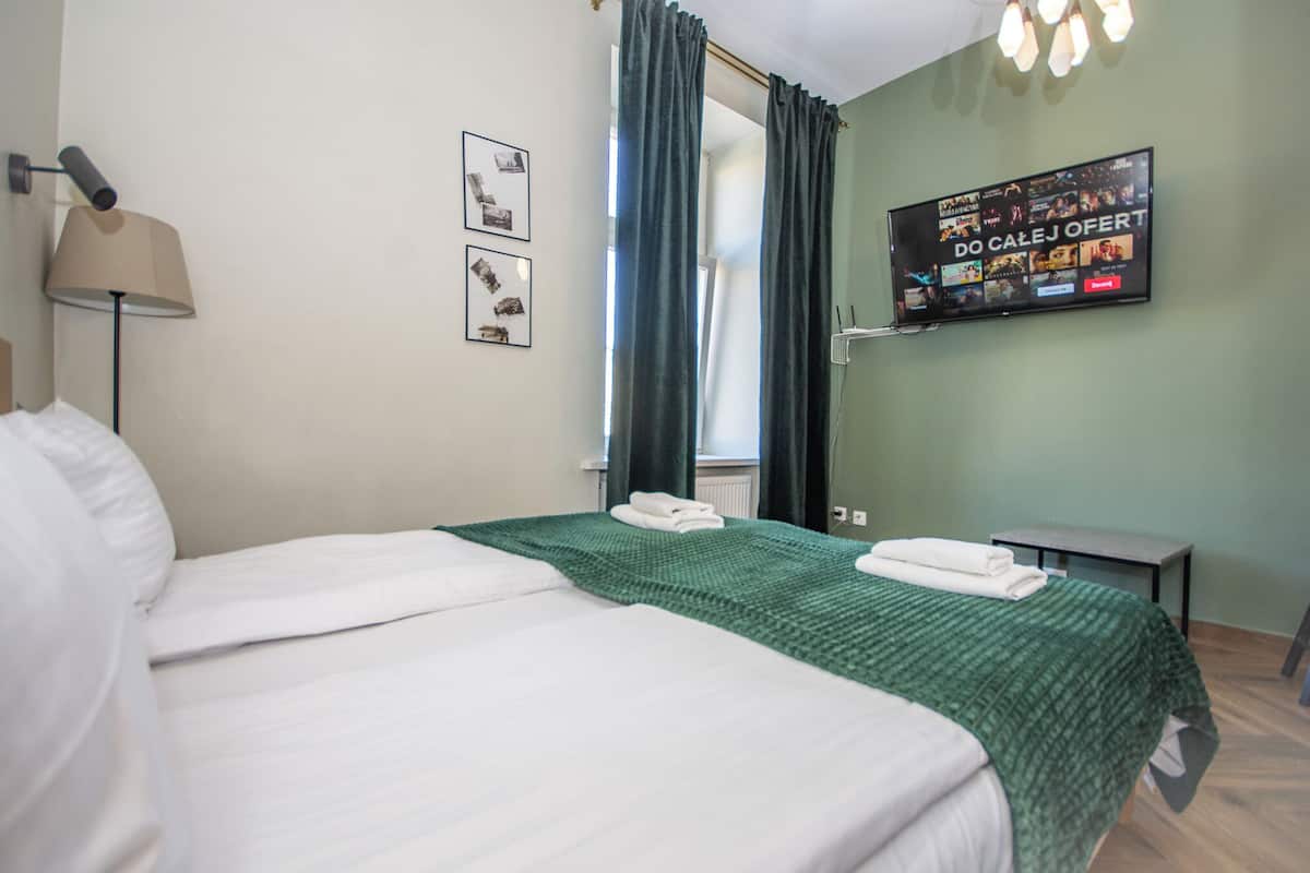 Quarto de hotel com cama, televisão na parede, luminária e janela com cortinas.