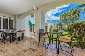 A covered patio with a table and chairs, a view of the garden, and a building with a white exterior and blue accents. - Palms At Wailea 301 1 Bedroom Condo (Kihei)