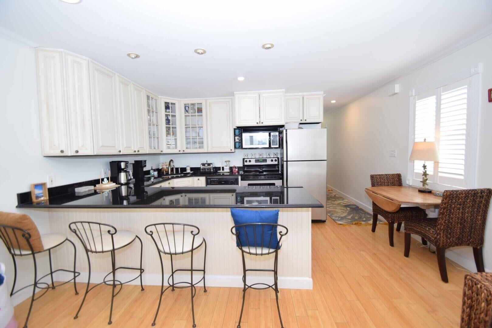 A modern kitchen with white cabinets, a central island with bar stools, a dining area with a table and chairs, and a refrigerator.