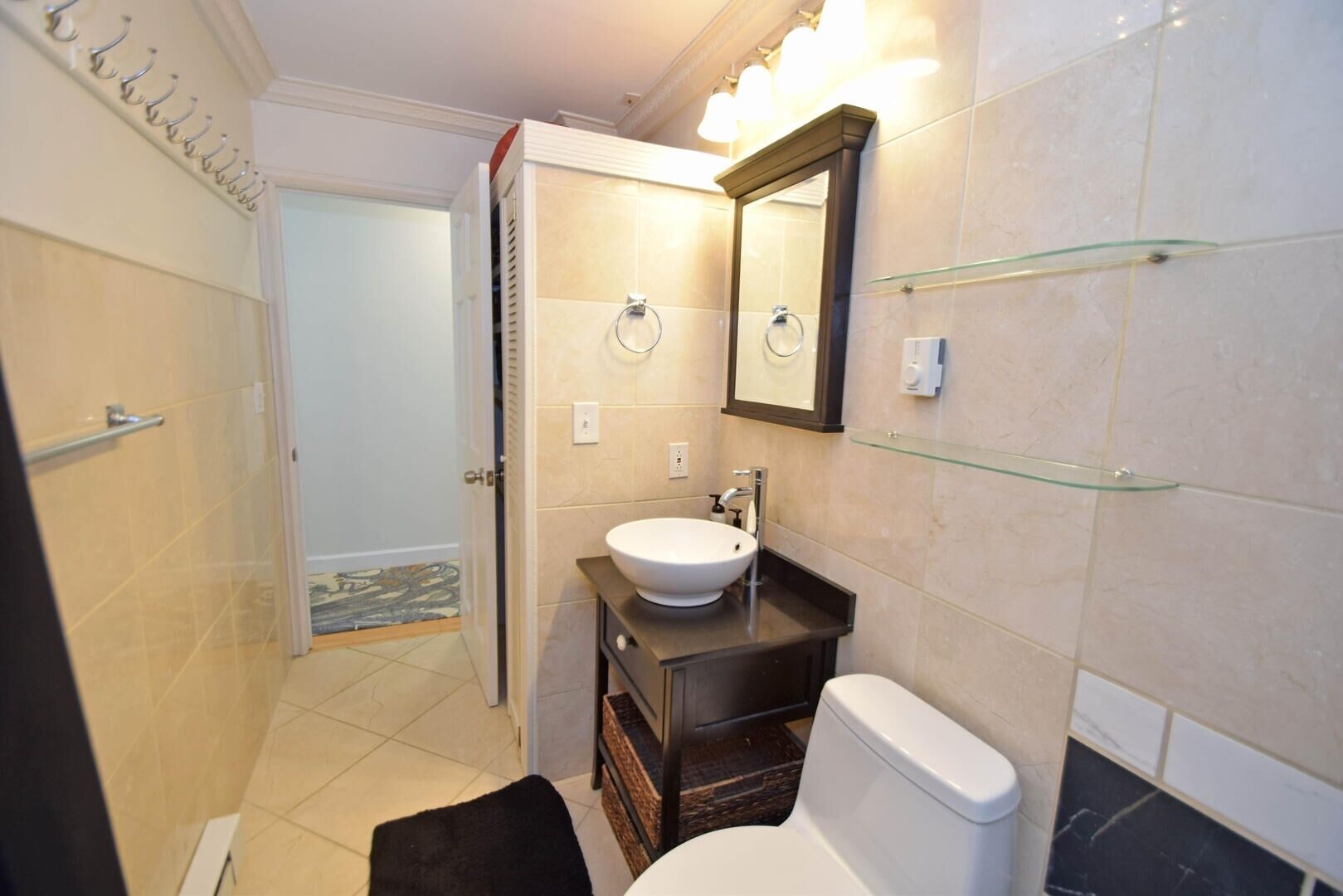 A bathroom with a white sink, dark countertop, glass shelves, a mirror, and a toilet.