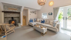 A living room with a fireplace, sofa, coffee table, wicker chair, and a large window. - Penbryn-Bach -  a self catering cottage that sleeps 10 guests in 4 bedrooms (Uwchmynydd)
