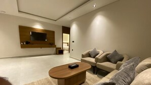 A modern living room with a flat-screen TV mounted on the wall, a wooden coffee table, and a beige sofa with grey cushions. - Onsen Hotel Al Shati (Dammam)
