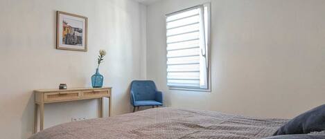 A bedroom with a bed, a chair, a desk, a picture on the wall, and a window with blinds.