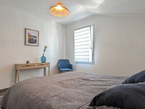 A bedroom with a bed, a chair, a desk, a picture on the wall, and a window with blinds. - Maison du Maine (Le Grand-Village-Plage)