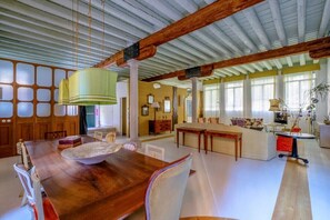 Une pièce spacieuse avec une table à manger en bois, des chaises, un luminaire suspendu et une grande fenêtre laissant entrer la lumière naturelle.