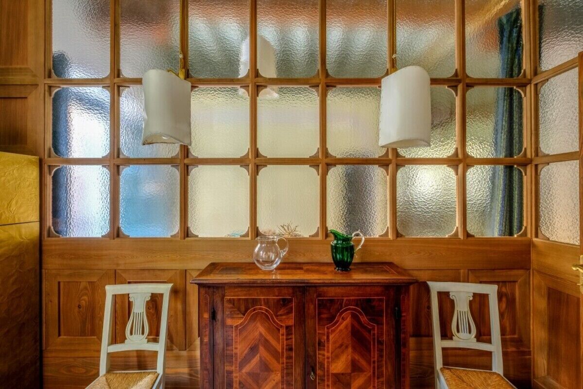 Une pièce en bois avec une grande fenêtre, deux chaises blanches, une table en bois et deux lampes fixées au mur.
