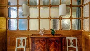 Ein Zimmer mit Holzboden, einem großen Fenster, zwei weißen Stühlen, einem Holztisch und zwei Wandleuchten.