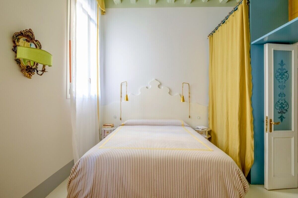 Un dormitorio con cama, lámparas de noche, mesita de noche, una puerta con panel decorativo y ventana con cortinas.