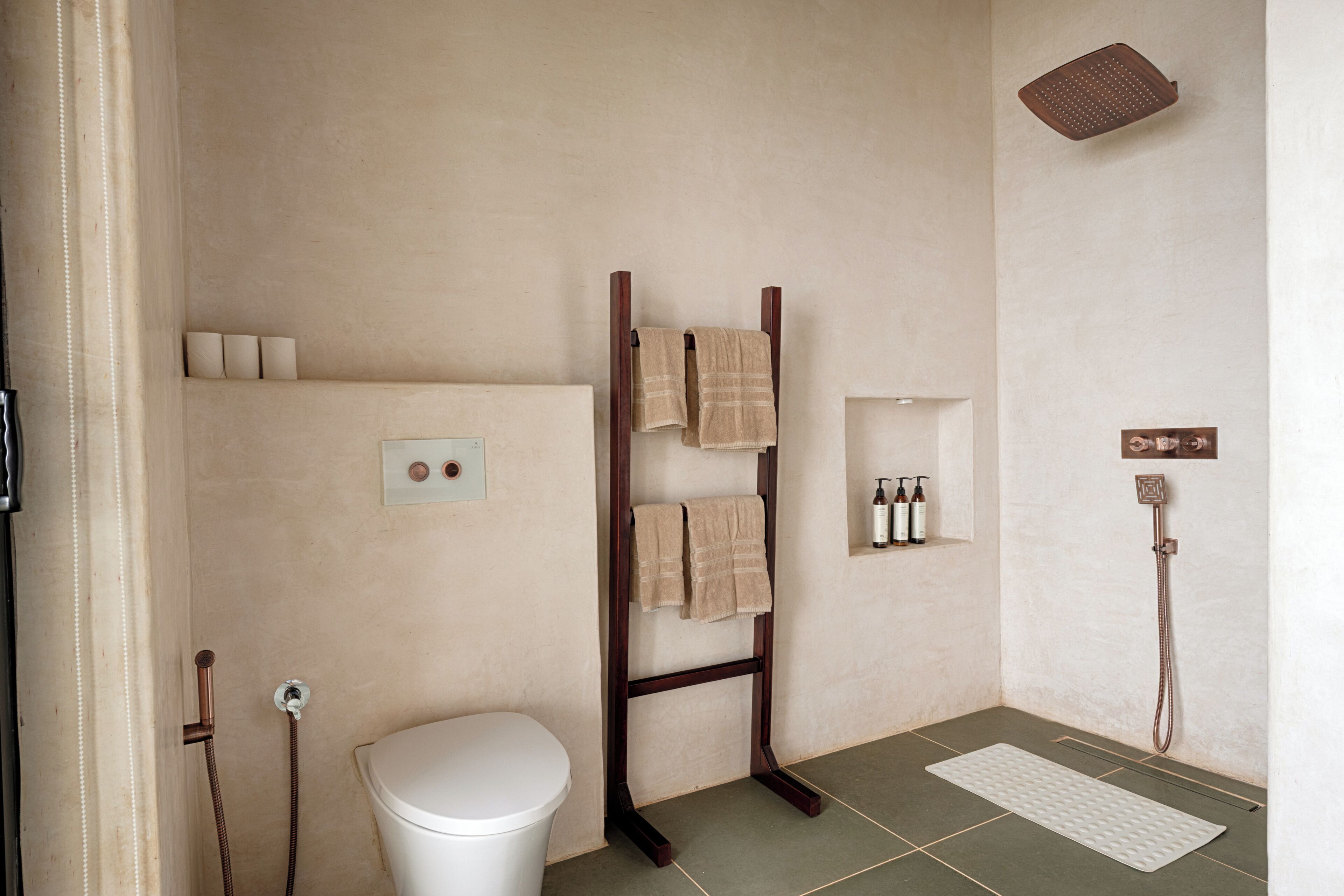 A bathroom with a white toilet, a wooden towel rack with towels, a shower, and a shelf with toiletries.