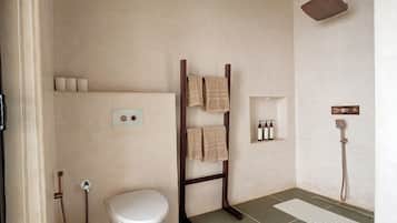 A bathroom with a white toilet, a wooden towel rack with towels, a shower, and a shelf with toiletries.
