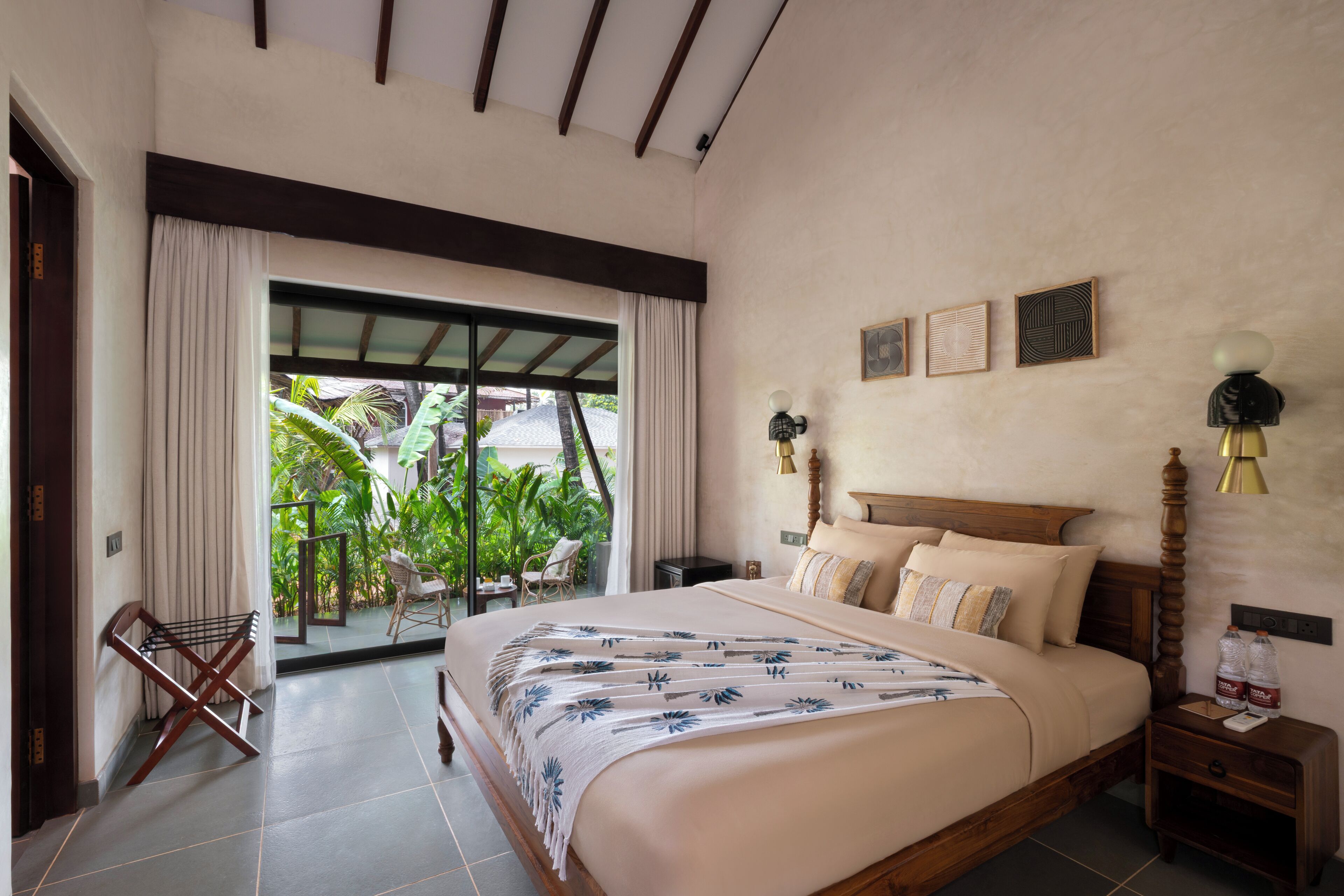 A bedroom with a large bed, a nightstand, a lamp, and a view of a tropical garden through a sliding door.