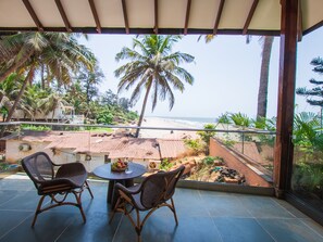 A balcony with wicker chairs and a table, overlooking a beach and palm trees. - The Earth-Maison Ocean Beach Resort (Ashvem)