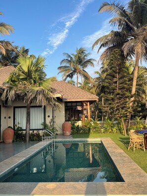 A pool with a clear blue sky, surrounded by palm trees and a building with a tiled roof. - The Earth-Maison Ocean Beach Resort (Ashvem)
