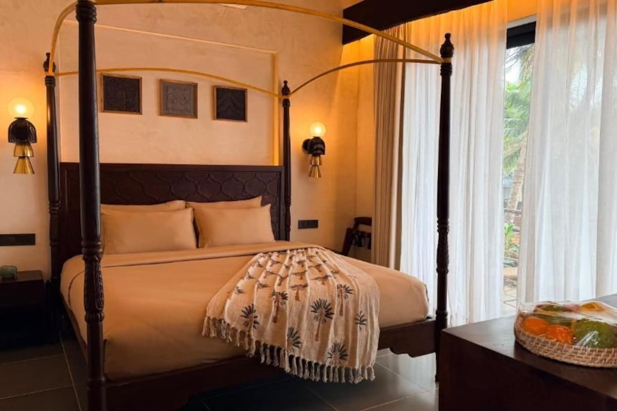 A four-poster bed with a canopy, a bedside table with a fruit basket, and a view of greenery outside.