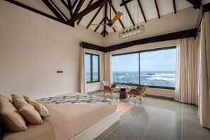 A bedroom with a large bed, a dining table with chairs, a ceiling fan, and a view of the ocean. - The Earth-Maison Ocean Beach Resort (Ashvem)