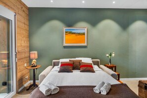 Ein Schlafzimmer mit einem Bett, Kissen, einem Nachttisch, einer Lampe, einem Gemälde und einer Vase mit Blumen.
