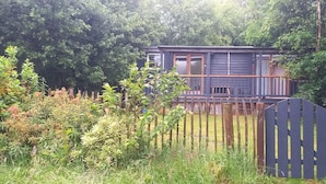 A cabin with a deck, surrounded by dense greenery and a wooden fence. - Sheperd's Hut, Sleeps 2 With Hot Tub and Garden (Glengarriff)