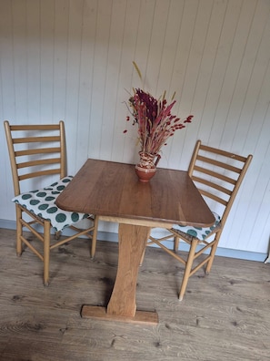 A wooden dining table with a vase of dried flowers, two wooden chairs with patterned cushions, and a wooden floor.