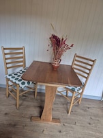 A wooden dining table with a vase of dried flowers, two wooden chairs with patterned cushions, and a wooden floor.