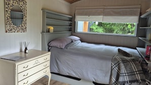 A bedroom with a bed, a desk with drawers, a mirror, shelves, and a window with a view of greenery. - Sheperd's Hut, Sleeps 2 With Hot Tub and Garden (Glengarriff)