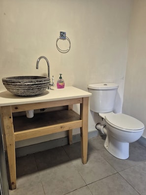 A bathroom with a wooden vanity, a round basin, a faucet, a toilet, and a soap dispenser.