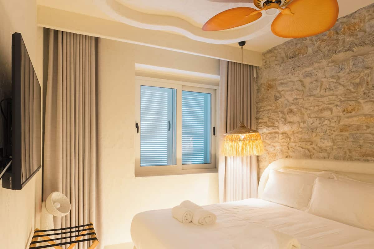 A bedroom with a bed, a window with shutters, a stone wall, a ceiling fan, and a TV mounted on the wall.