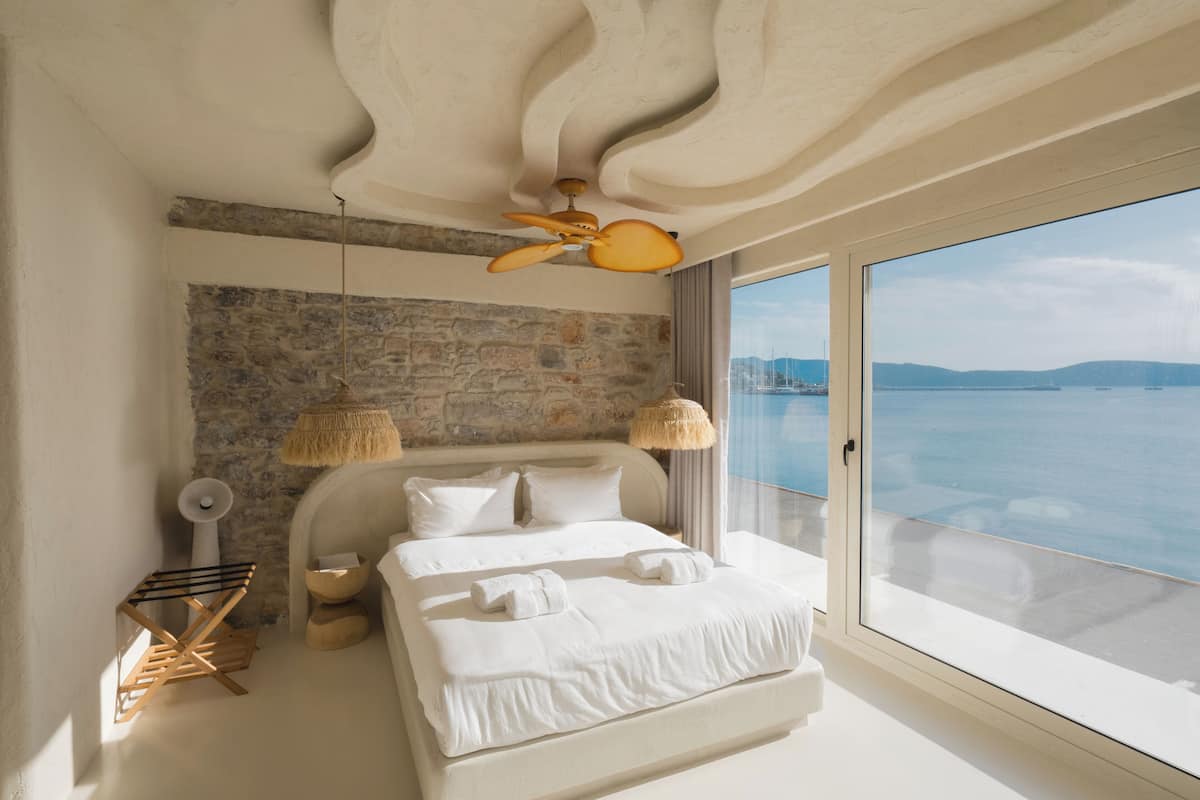 A bedroom with a stone wall, a large bed, a ceiling fan, and a view of the sea.
