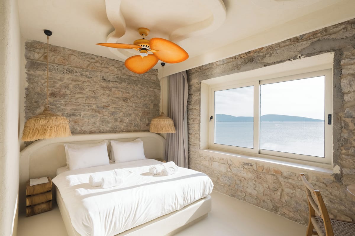 A bedroom with a stone wall, a bed with white linens, a ceiling fan, and a window with a view of the sea.