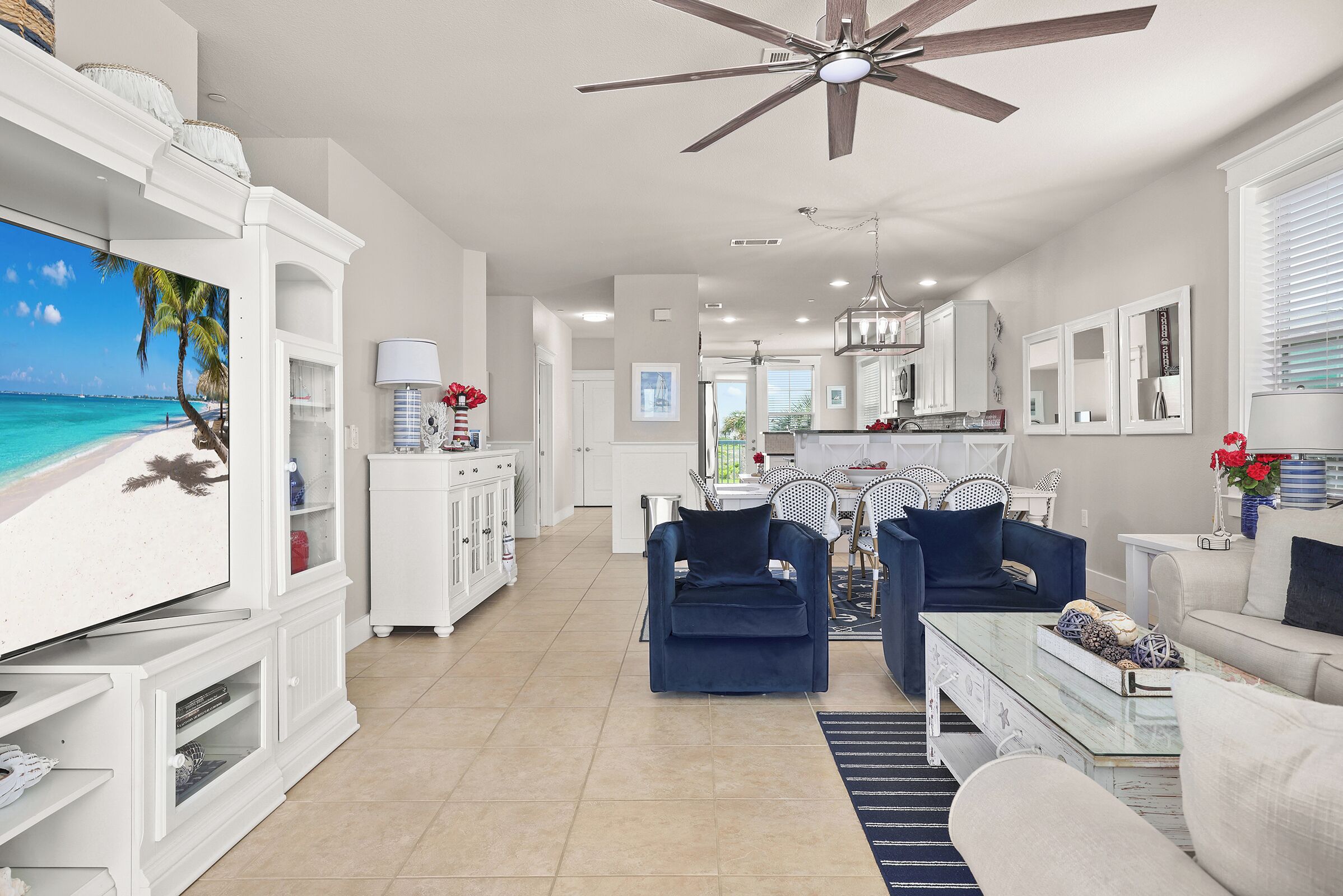 A spacious living area with a ceiling fan, a television displaying a beach scene, and a dining area with chairs and a table.