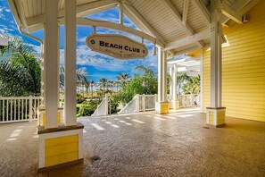 Ein überdachter Außenbereich mit einem Schild, auf dem 'Beach Club' steht, und Palmen im Hintergrund.