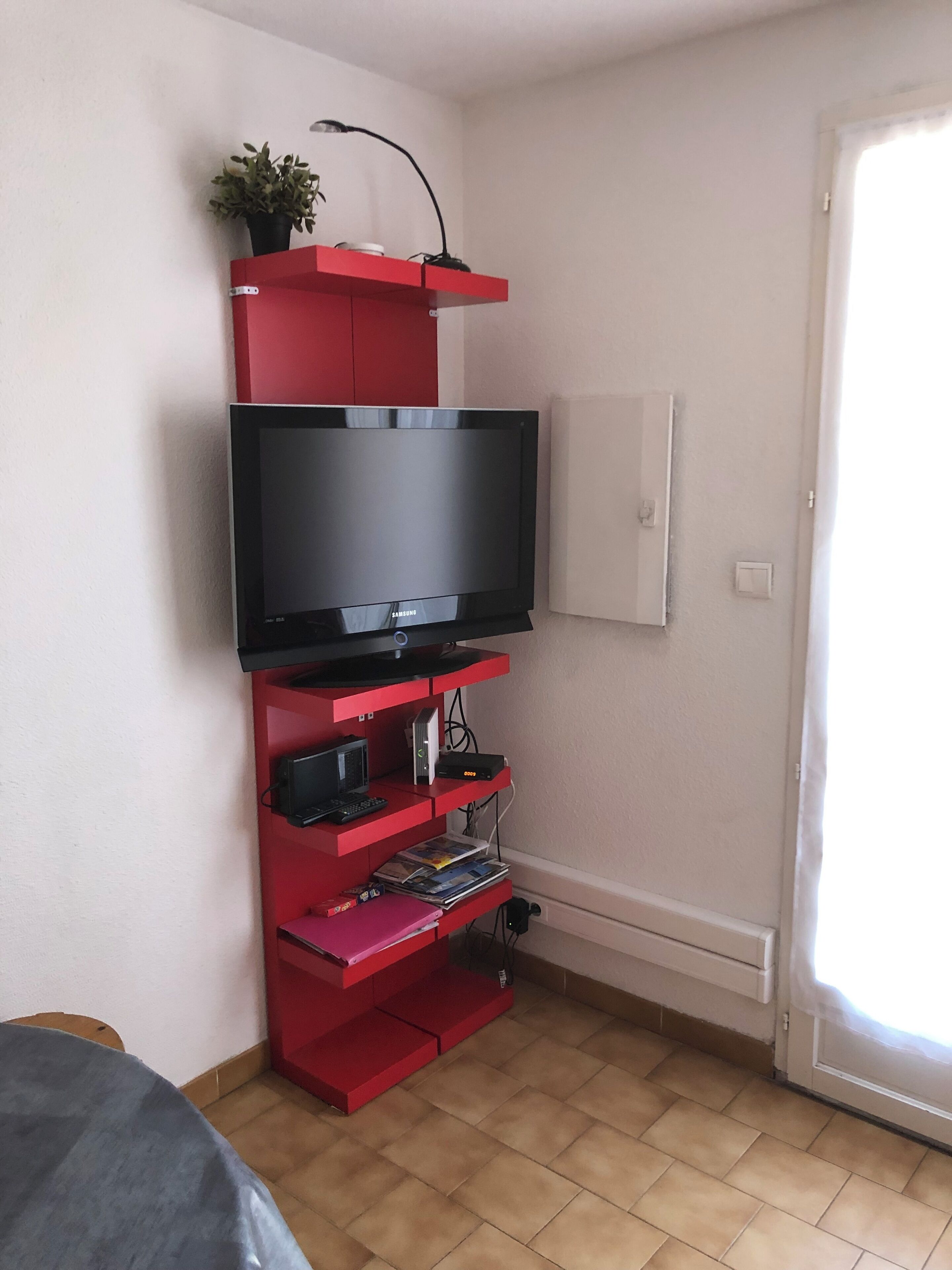 Une pièce comprenant un meuble de rangement rouge pour les appareils multimédias, un téléviseur à écran plat, un petit bureau et une porte.