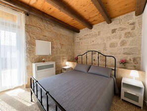 Ein Schlafzimmer mit Steinwand, Holzboden, einem Bett mit Metallgestell, einem Nachttisch mit Lampe und einer Kommode.