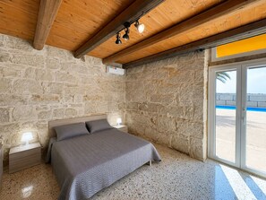 A bedroom with a stone wall, a bed with a grey bedspread, a nightstand with a lamp, and a window with a view of the beach. - Stunning private villa with WIFI, private pool, A/C, TV, patio, pets allowed and panoramic view (Santa Croce Camerina)
