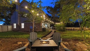 Un jardin avec un foyer intégré dans une table et des fauteuils Adirondack, le tout entouré d’une terrasse en bois et illuminé par des guirlandes lumineuses.