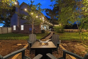 Ein Garten mit Feuerstelle, Adirondack-Stühlen und einer Holzterrasse, umgeben von Lichterketten.